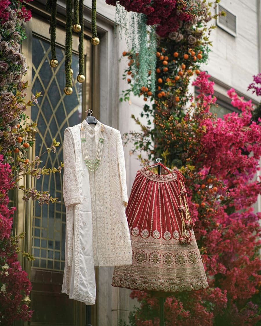 A cream sherwani and a red bridal lehenga hanging under a floral arch